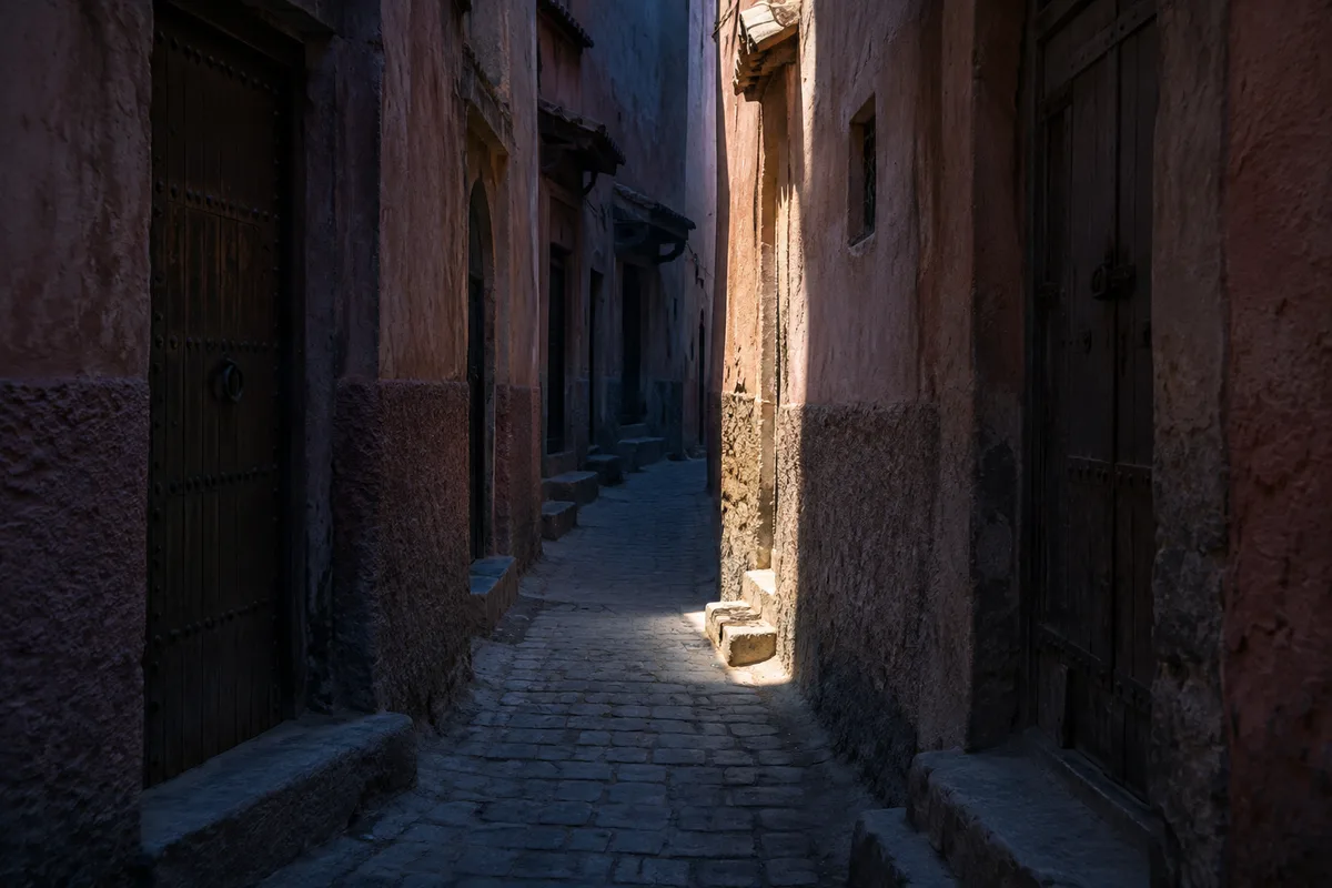 Ruelle étroite aux murs de chaux rosée traversée par un unique faisceau de lumière tombant entre deux bâtiments